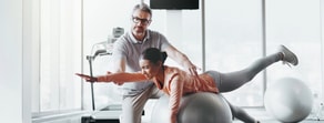 A trainer helps a woman with a balance exercise on a stability ball, integrating hybrid care, in front of treadmills and large windows.