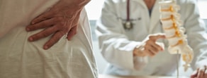 A patient holds their waist in pain as a doctor points to a spine model alongside a medbridge display.