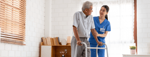 A nurse helps an elderly man with a walker in a well-lit room with white brick walls, bookshelves, and window blinds.