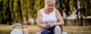 A woman in a white tank top and purple pants sits on the grass, adjusting her shoelaces next to a kettlebell and water bottle, with tall trees behind her.