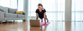 A woman planks on a pink yoga mat using Medbridge guidance on her laptop.