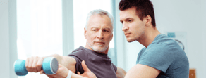 In a gym, an older man lifts a dumbbell with support from a younger coach, part of medbridge healthcare training.