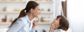 A woman and child smile at each other, touching their chins, in a room with background shelves.