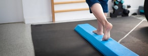 A person balancing barefoot on a blue foam beam in a medbridge fitness studio.