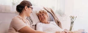 A caregiver with glasses smiles warmly at an elderly woman in bed, emphasizing the role of Medbridge's digital tools in fostering meaningful connections.