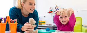 A woman at a table with a child in a pink shirt crafts colorful items, holding a teddy bear.