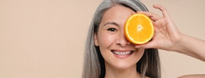 A smiling woman with long gray hair holds half an orange over one eye against a beige background.