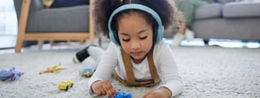 A child wearing headphones plays with toy cars on a carpet as adults sit on a sofa in the background.