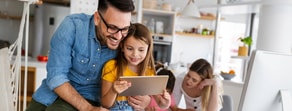 A man and girl smile at a tablet with a woman and child in the kitchen behind them, using medbridge.
