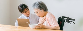 An elderly woman in a wheelchair coughs while a caregiver with a clipboard watches her at a table, illustrating hybrid care with medbridge.