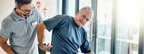A physical therapist helps an older man with back exercises in a bright medbridge office.