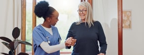 A healthcare worker in scrubs helps a smiling older woman in a bright room, illustrating medbridge hybrid care.