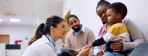 A healthcare worker talks to a boy on his mother's lap as another adult takes notes in a clinic, emphasizing medbridge solutions.
