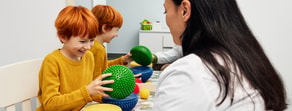 Two red-haired children play with sensory balls at a table as a dark-haired adult supervises, illustrating medbridge activities.