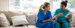Two healthcare professionals in scrubs sit on a couch, viewing digital medbridge solutions on a phone, notebook, and tablet.