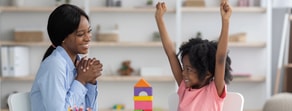 A woman smiles at an excited child with raised arms at a table of colorful blocks, reflecting Medbridge healthcare education.