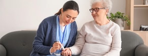 A healthcare worker checks an elderly woman's blood sugar with a glucose meter on a sofa, illustrating medbridge technology.