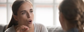 A woman gestures as she talks about healthcare education with someone indoors, a window visible behind them.