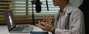 A person in a white coat and stethoscope records a healthcare podcast at a desk with a laptop and notebook.