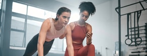 Two women in a gym, one exercising and one timing her with a stopwatch, with gym equipment and medbridge-style training implied.