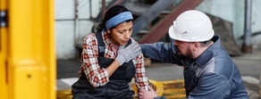 A worker sits holding her shoulder as a coworker in a hard hat kneels to help, highlighting medbridge healthcare training.
