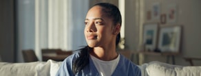 A woman in blue scrubs sits on a couch, looking thoughtful, with sunlight streaming in—medbridge software on her mind.