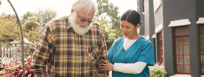 A nurse with Medbridge software helps an elderly man with a white beard walk outside near a building.