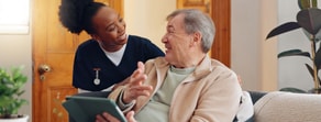 A nurse in scrubs smiles at an older man holding a tablet, with plants and a wooden door shown in a medbridge healthcare setting.