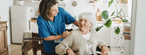 A caregiver in blue scrubs helps an elderly woman in a wheelchair in a bright kitchen, showing medbridge hybrid care.