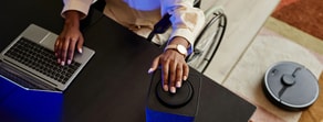 A wheelchair user accesses medbridge on a laptop and adjusts a smart speaker as a robotic vacuum moves nearby.