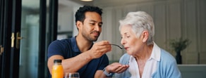 A young man helps feed an elderly woman at a dining table, with a glass of orange juice in front, for medbridge healthcare.