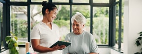 A caregiver with healthcare training helps an older woman in a bright room with large windows and green plants, holding a tablet.