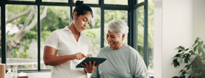 A young woman shows an older woman patient care info on medbridge software in a bright, plant-filled room with large windows.