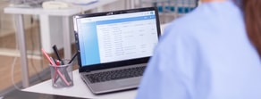 A person in scrubs sits at a desk viewing medical records on a laptop, with a pen holder nearby.