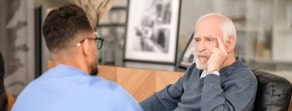 A young man in scrubs talks to an older man seated indoors, illustrating medbridge’s role in healthcare training.