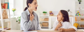 A woman and a girl touch their throats and vocalize together at a desk in a bright, tidy medbridge therapy room.
