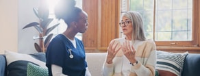 A nurse in blue scrubs listens to an older woman with gray hair and glasses talking on a sofa.