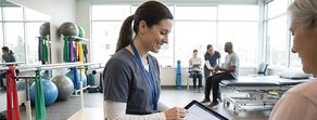 A healthcare worker uses a tablet with medbridge software while speaking to a patient in a rehab clinic.