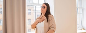 A woman with glasses stands indoors by a window, eyes closed and hand on chest, looking calm and thoughtful—reflecting the mindful approach promoted by medbridge.
