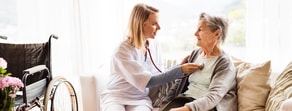 A healthcare professional uses a stethoscope on an elderly woman near a wheelchair.