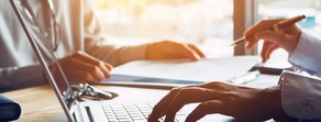 Two individuals work at a desk with a laptop and documents, focusing on medbridge software.