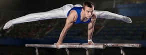 In a stadium, a gymnast in a blue top performs a split handstand on parallel bars, exemplifying precision akin to medbridge training.