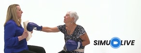 An older woman in boxing gloves punches a board held by a seated woman, with the SimuLive logo in the corner.