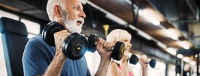 An older man and a woman exercise with weights in a well-lit gym for medbridge training.