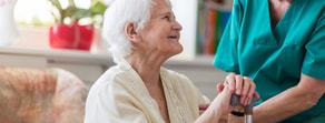 An elderly woman using a walking stick is helped by a caregiver in green, highlighting medbridge's digital healthcare in a cozy room.