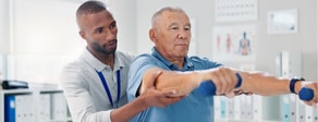 A physical therapist uses medbridge to help an older man lift dumbbells during rehab in a clinic.