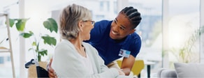 A caregiver in a blue shirt talks with an older woman in a wheelchair in a bright room, showing medbridge hybrid care.