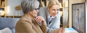 Two women smile at a computer showing a digital healthcare visit in a cozy home, using medbridge.