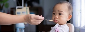 A baby in a high chair looks hesitant as an adult offers food with a spoon.