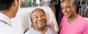 A patient in a hospital bed smiles at a healthcare provider, with another woman smiling beside him, showcasing modern digital healthcare advancements by medbridge.